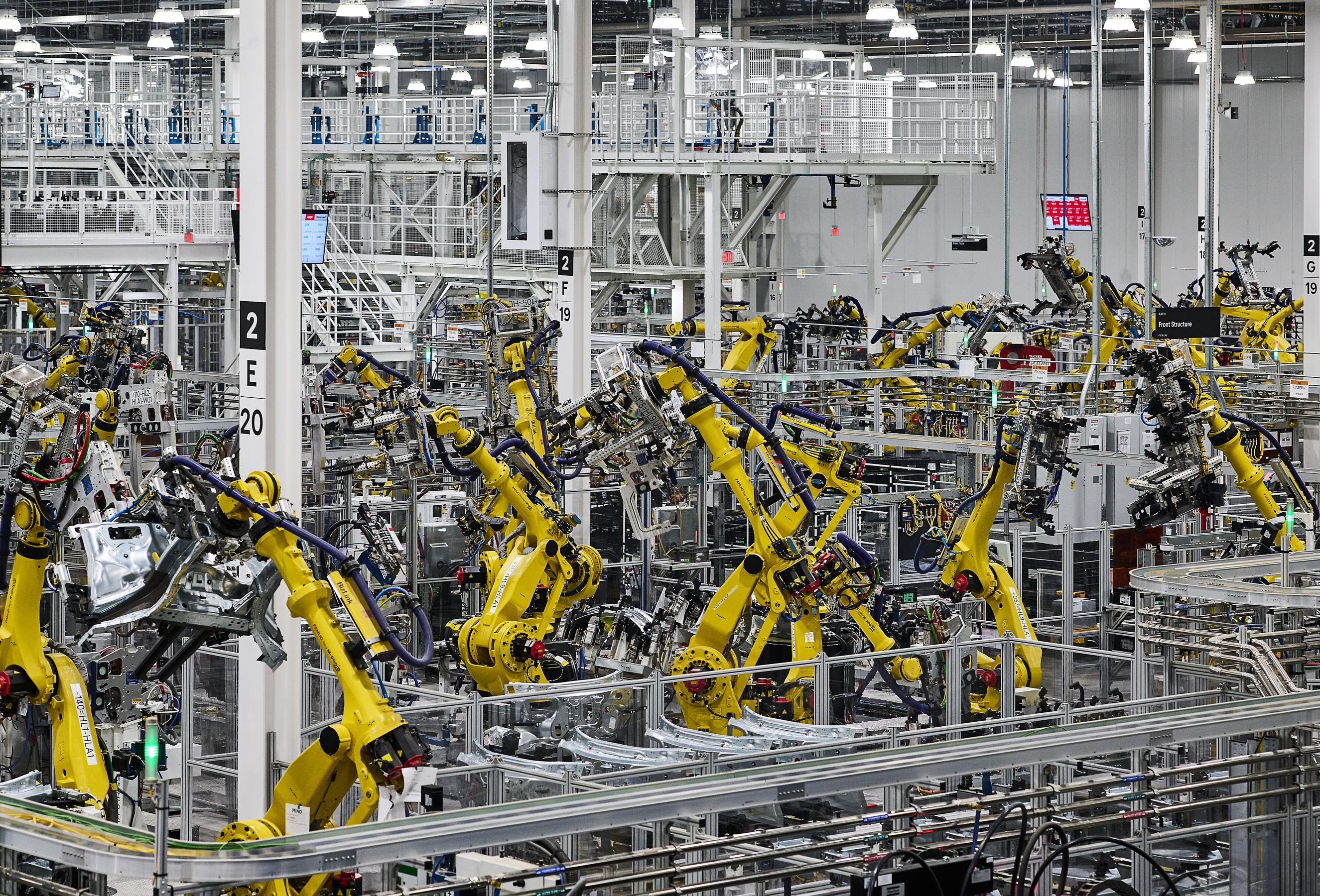 A wide and high view of the factory floor where yellow machine arms are seen lifting and working on unspecified car parts.