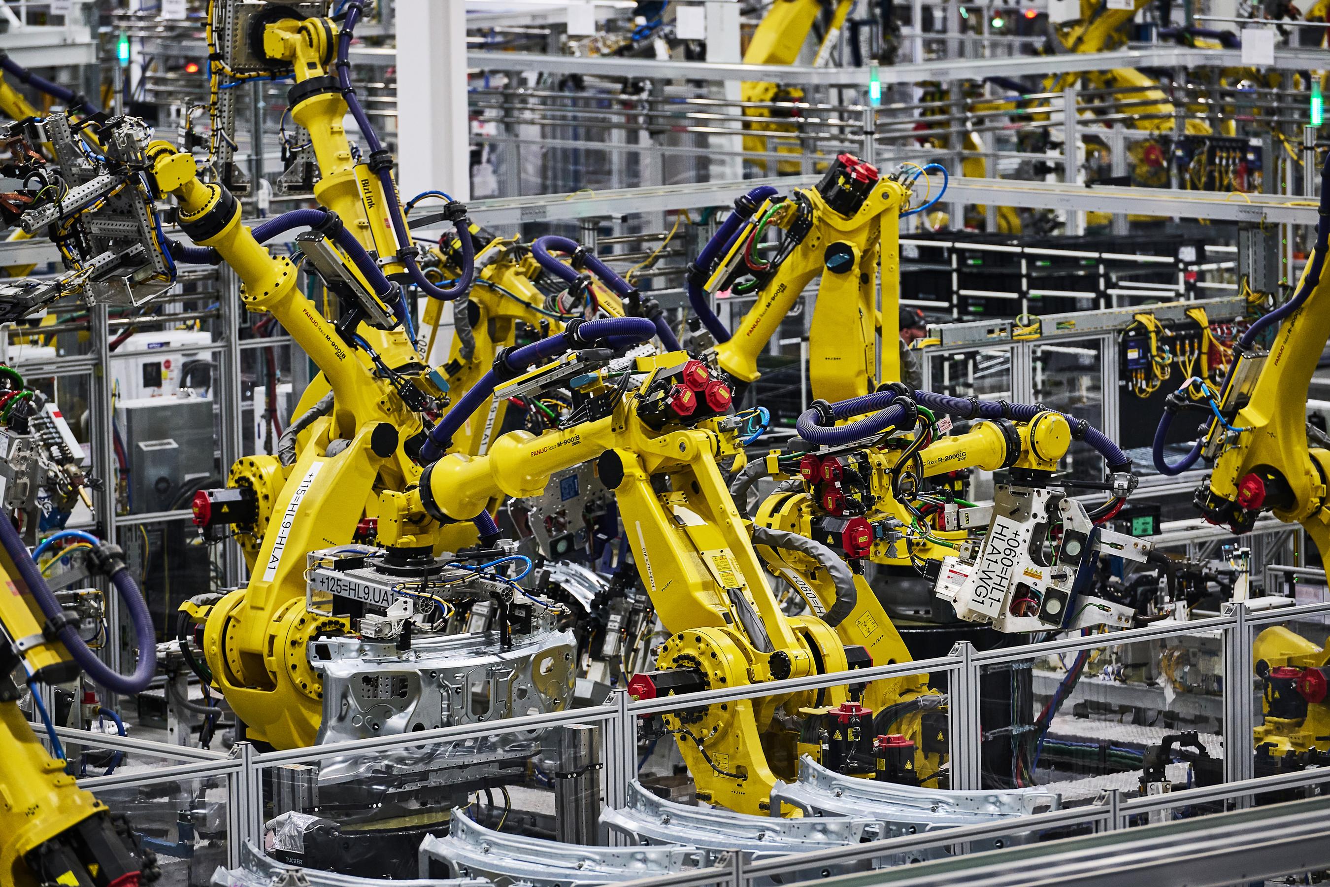 A cluster of yellow machine arms working on unspecified silver car parts on the Rivian factory floor.