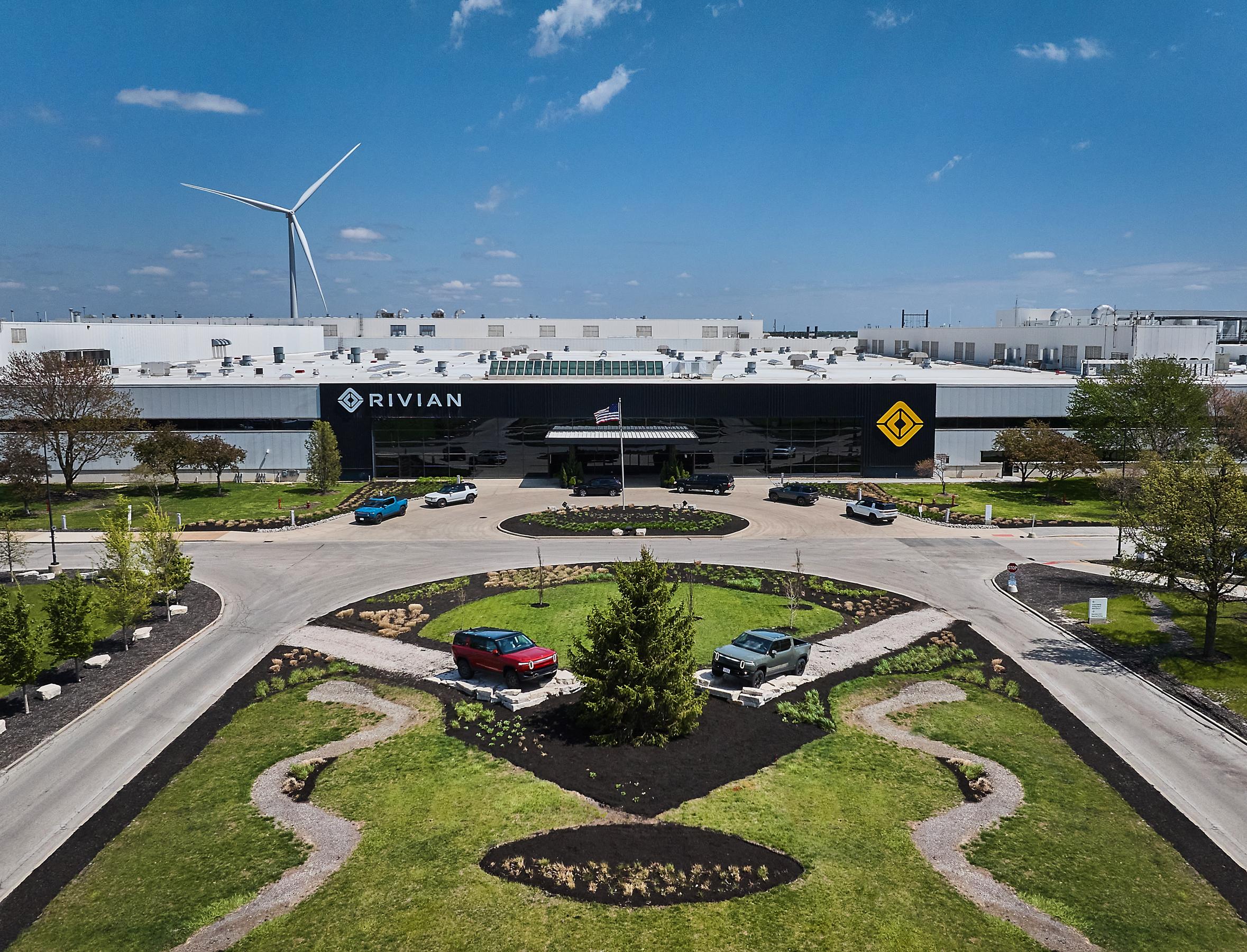 A drone shot of the entrance of the Rivian factory. There’s a manicured grassy area with two vehicles displayed along the road leading to the entrance of the enormous Rivian building. It’s a bright sunny day with a blue sky overhead.