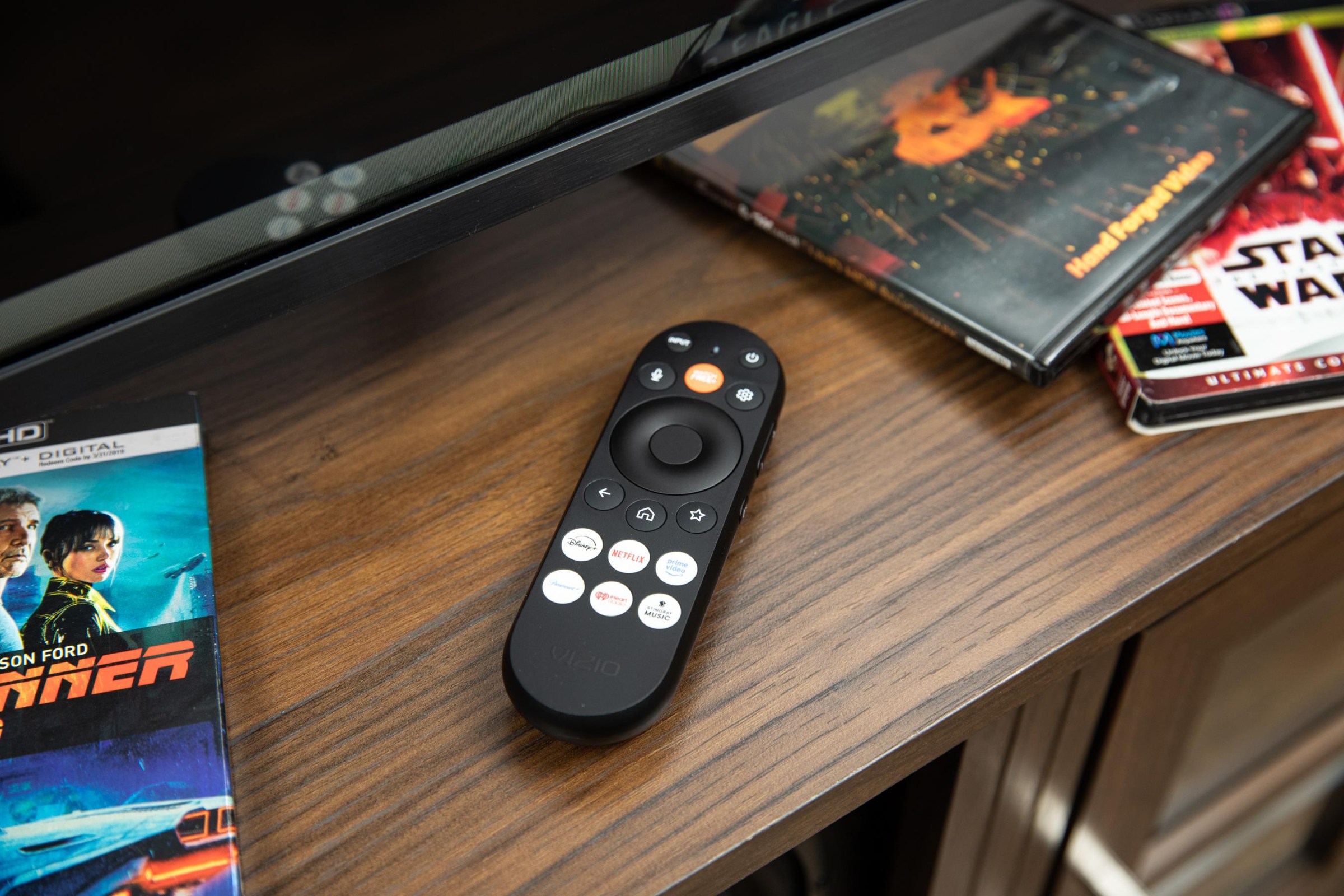 The remote for the Vizio Mini LED Quantum TV on a wooden credenza with Blu-ray disc cases around it.