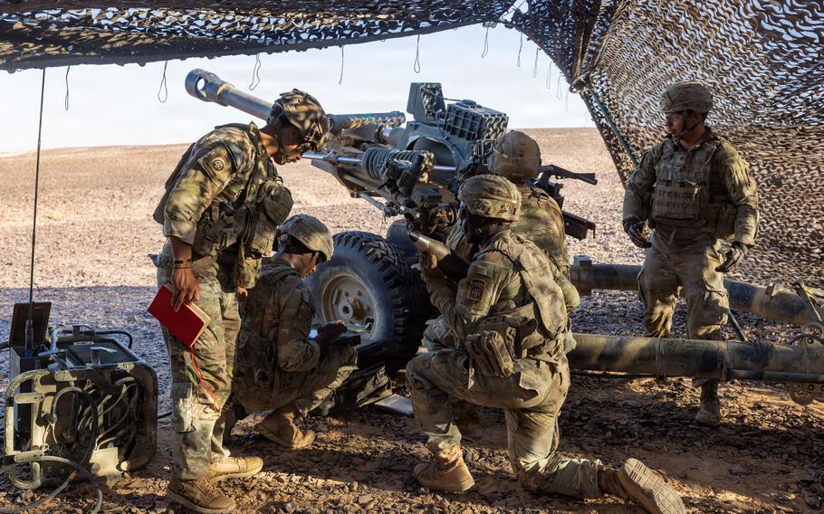 Soldiers kneel around an artillery piece under camouflage netting in the desert.