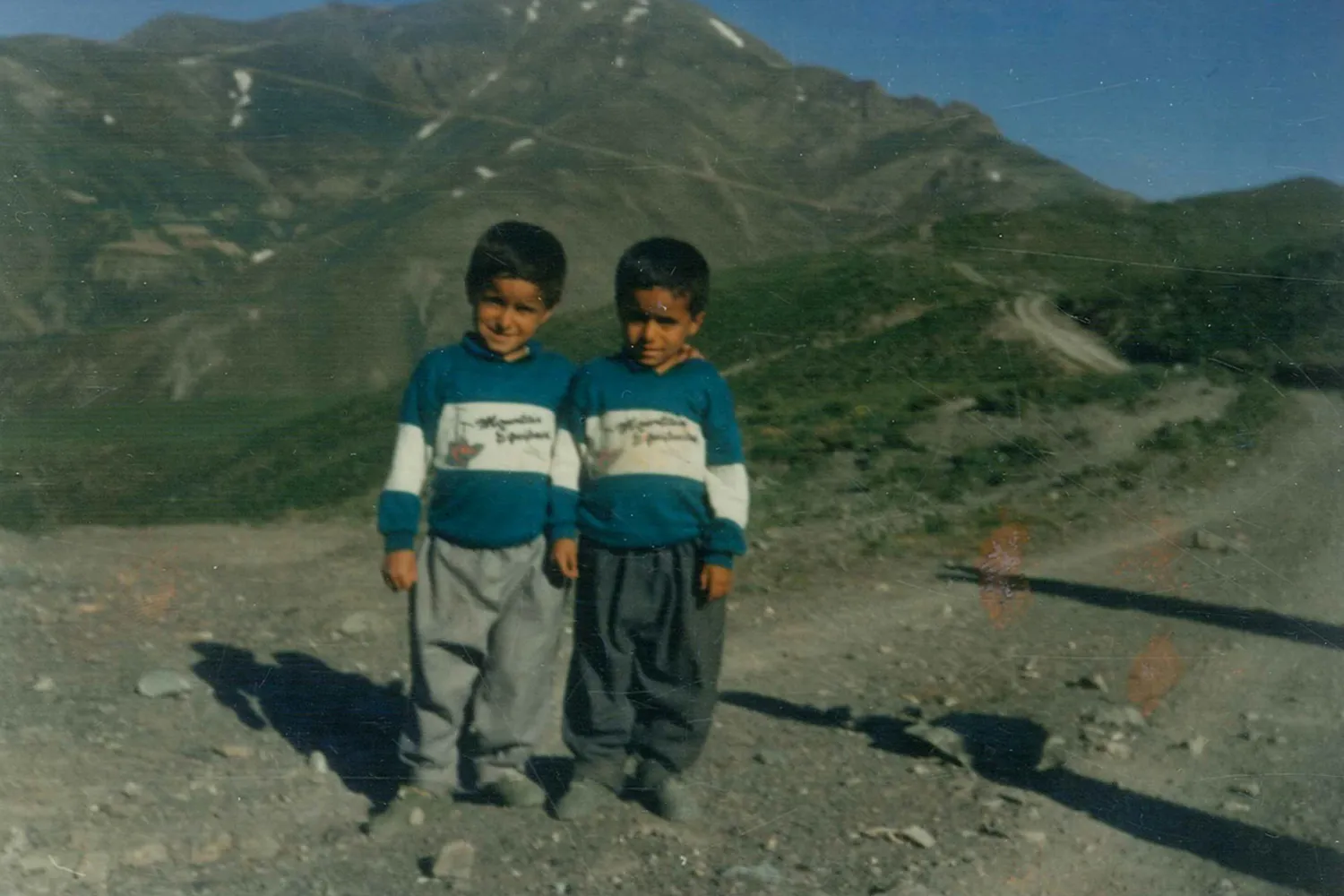 A slightly grainy, older outdoor photograph of two young boys standing side by side on a dirt path. They are wearing matching blue and white long-sleeved sweatshirts and baggy trousers. In the background, a large green and rocky mountain rises under a blue sky, with a winding path visible on the hillside to the right.