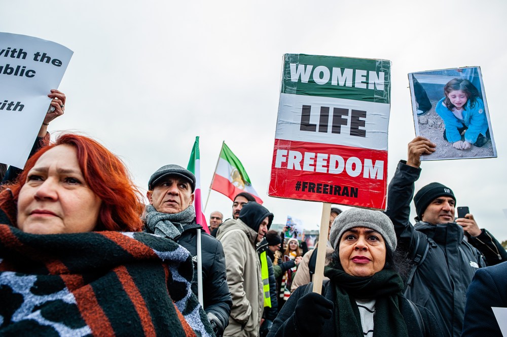 A crowd at an outdoor rally under an overcast sky. In the foreground, two women look toward the left of the frame. Behind them, people hold up various signs, including a prominent central sign painted with green, white, and red stripes that reads "WOMEN LIFE FREEDOM #FREEIRAN" in bold block letters. To the right, a person holds aloft a framed photograph of a young, crying child. Multiple flags with green, white, and red horizontal stripes are held up in the background.