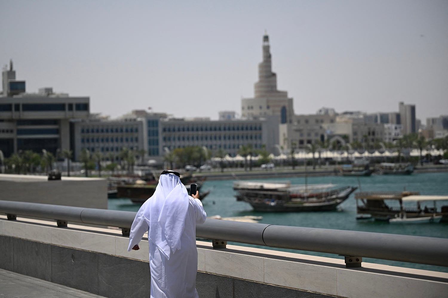 A person wearing a traditional white thobe and ghutra stands along a waterfront promenade holding a smartphone, with traditional wooden boats in the water and a city skyline featuring a distinctive spiral tower in the background.