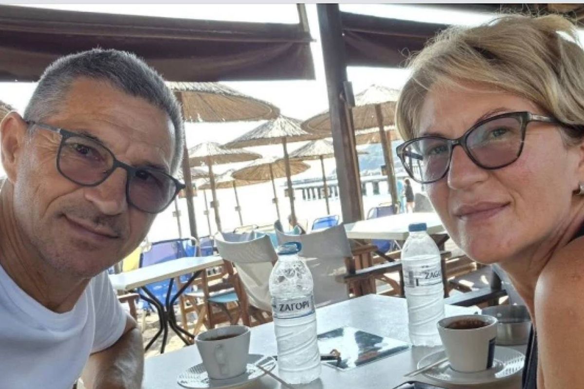 Two individuals are seated together at an outdoor table, possibly in a café setting. The man is wearing glasses and a white shirt, while the woman is wearing sunglasses and a sleeveless top. In front of them, there is a table arrangement featuring cups, bottles, and other items. The backdrop includes an umbrella and an open-air environment.