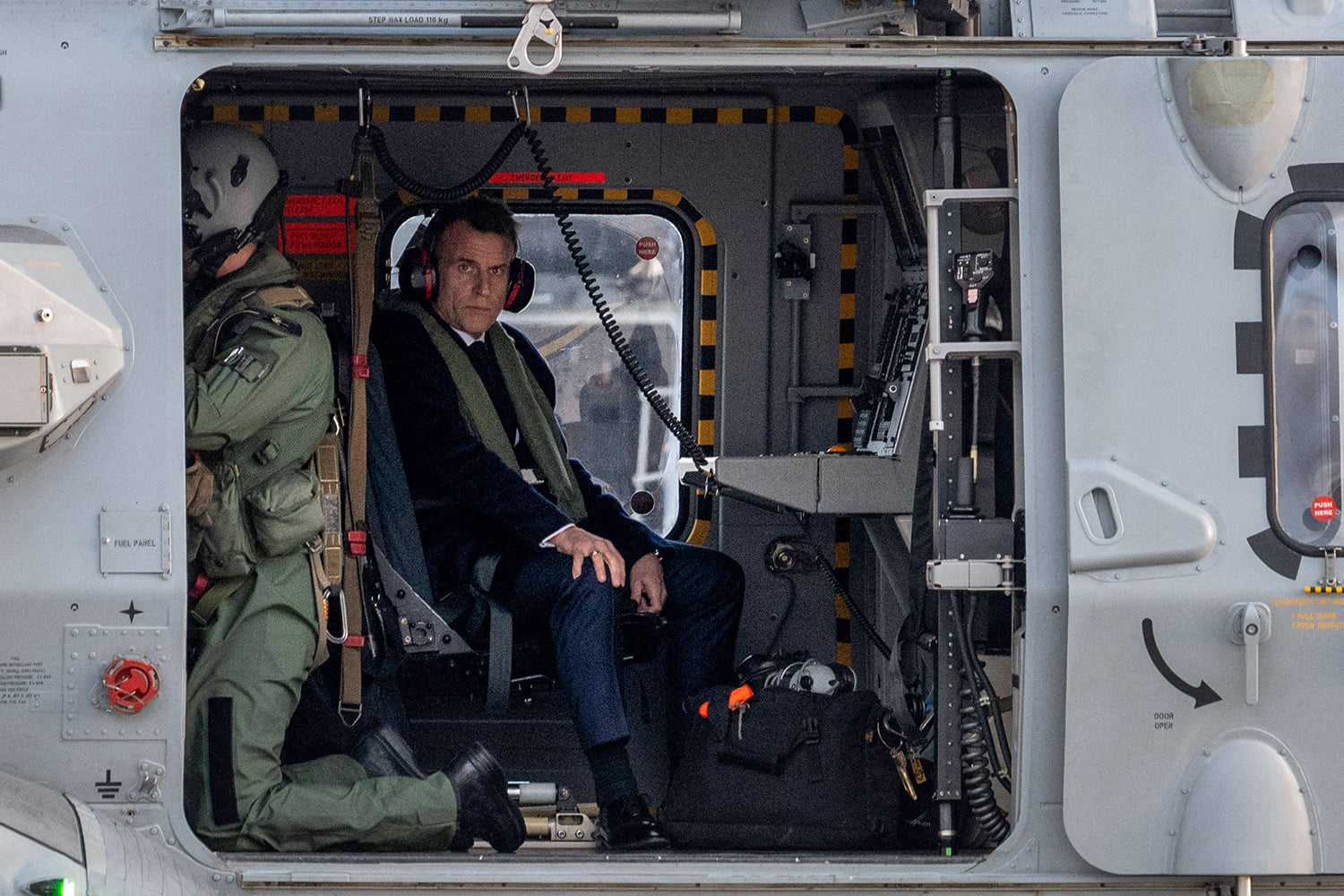 A man in a suit and noise-canceling headphones sits inside the open side door of a military helicopter next to a crew member wearing flight gear.
