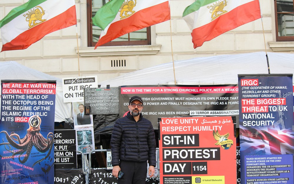 A man stands outdoors in front of a white tent structure decorated with numerous protest banners and signs. He has short dark hair, a beard, and wears glasses, a black baseball cap, and a black puffer jacket. Behind him, three flags with green, white, and red stripes and a central golden lion emblem fly against a light-colored stone building. The signs surrounding him feature text regarding the IRGC, national security, and a prominent red sign that reads "SIT-IN PROTEST DAY 1154."