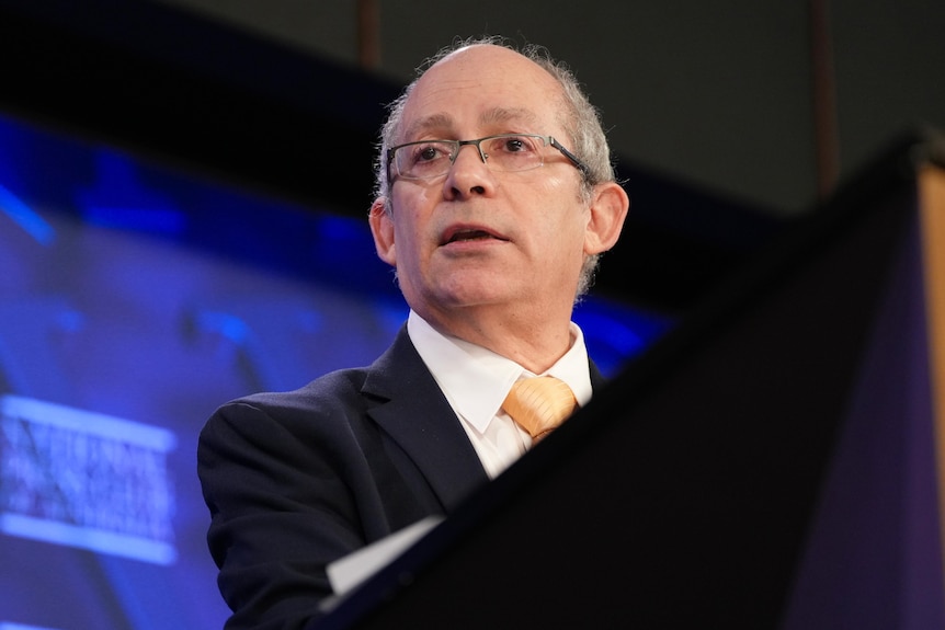 A white man with glasses speaks at a lectern in front of a blue background