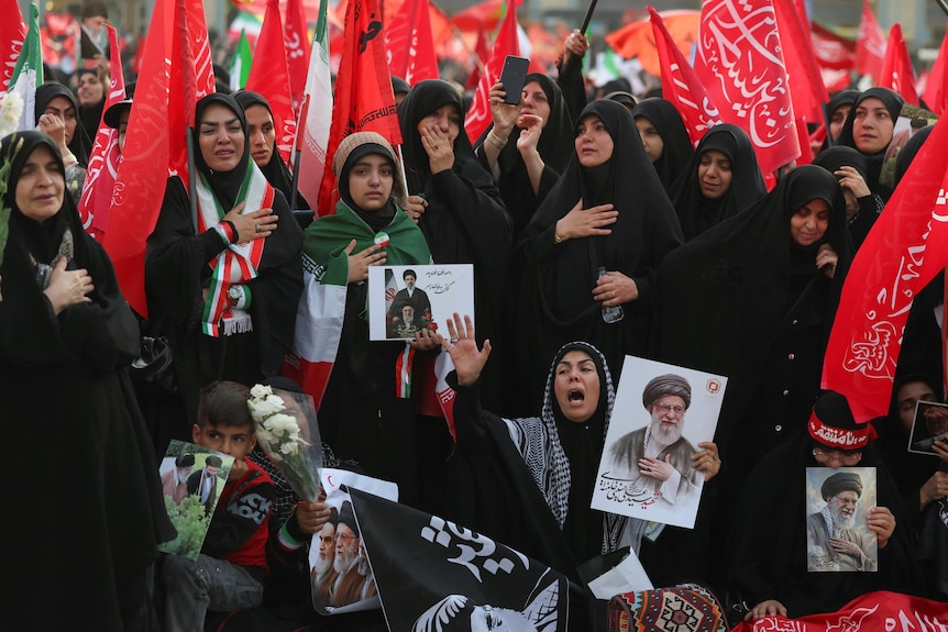 A crowd of women wearing black, draped with Iranian flags, mourning and holding Ali Khamenei posters.