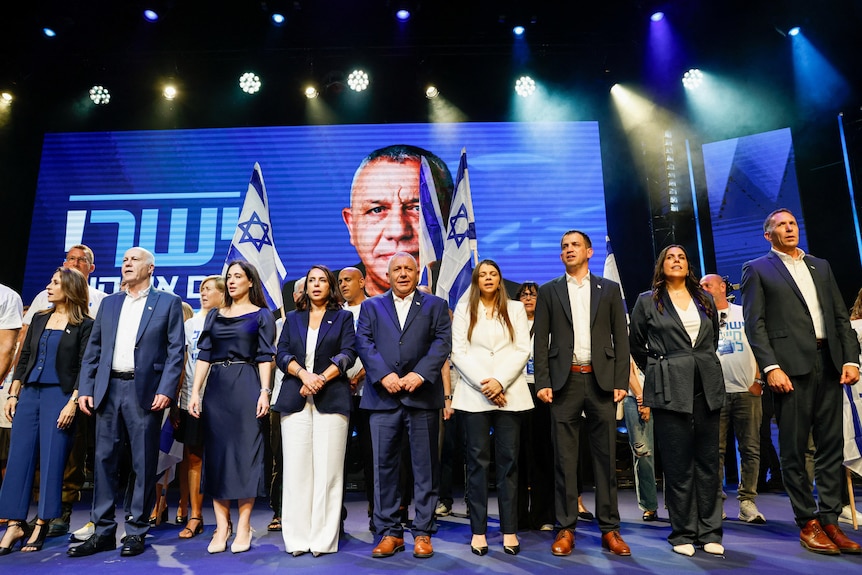 A line of men and women in suits and business attire signing in front of a large blue screen with the face of a man.