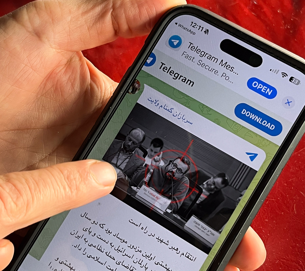 A close-up view of a person holding a smartphone against a red background. The phone screen shows a Telegram channel message featuring a black-and-white photograph of a man speaking at a panel discussion. A red digital crosshair or target symbol is overlaid directly onto the man's face in the photo. Below the image on the screen, text written in the Persian script is visible.