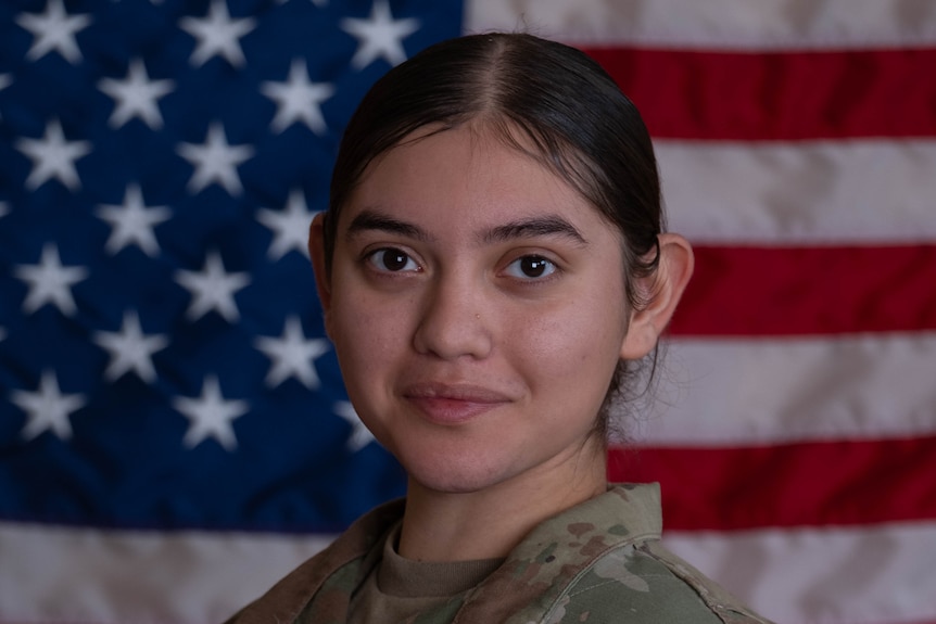 A young female soldier in army greens in front of a US flag.