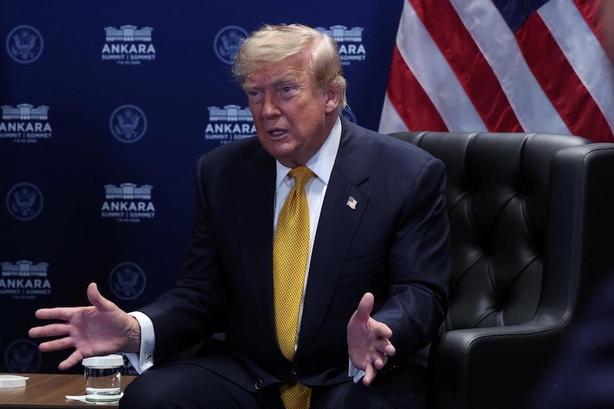 US president trump sits in a chair in front of blue background.