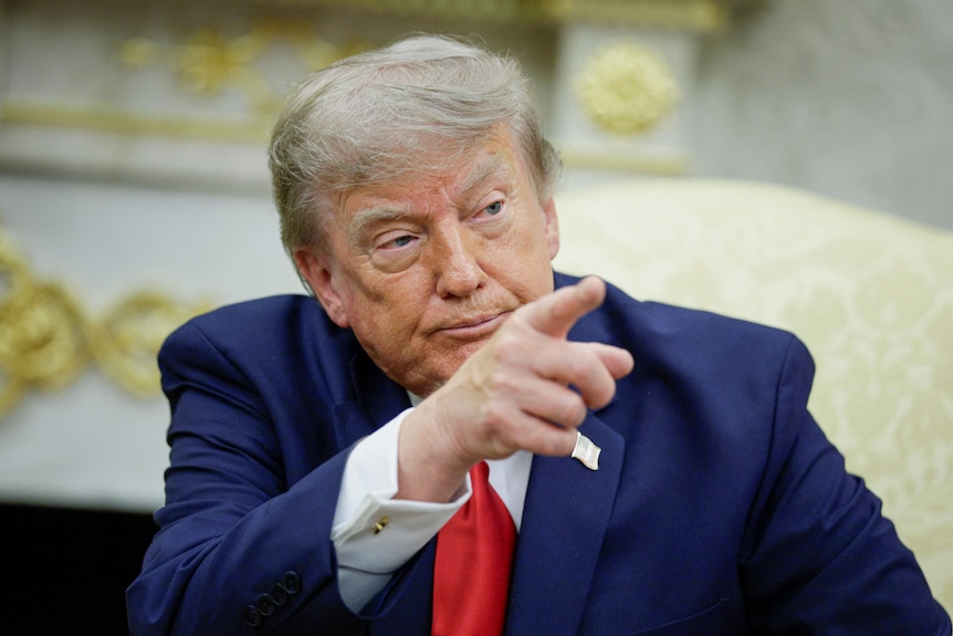Trump points to someone off-camera while sitting in the Oval Office.