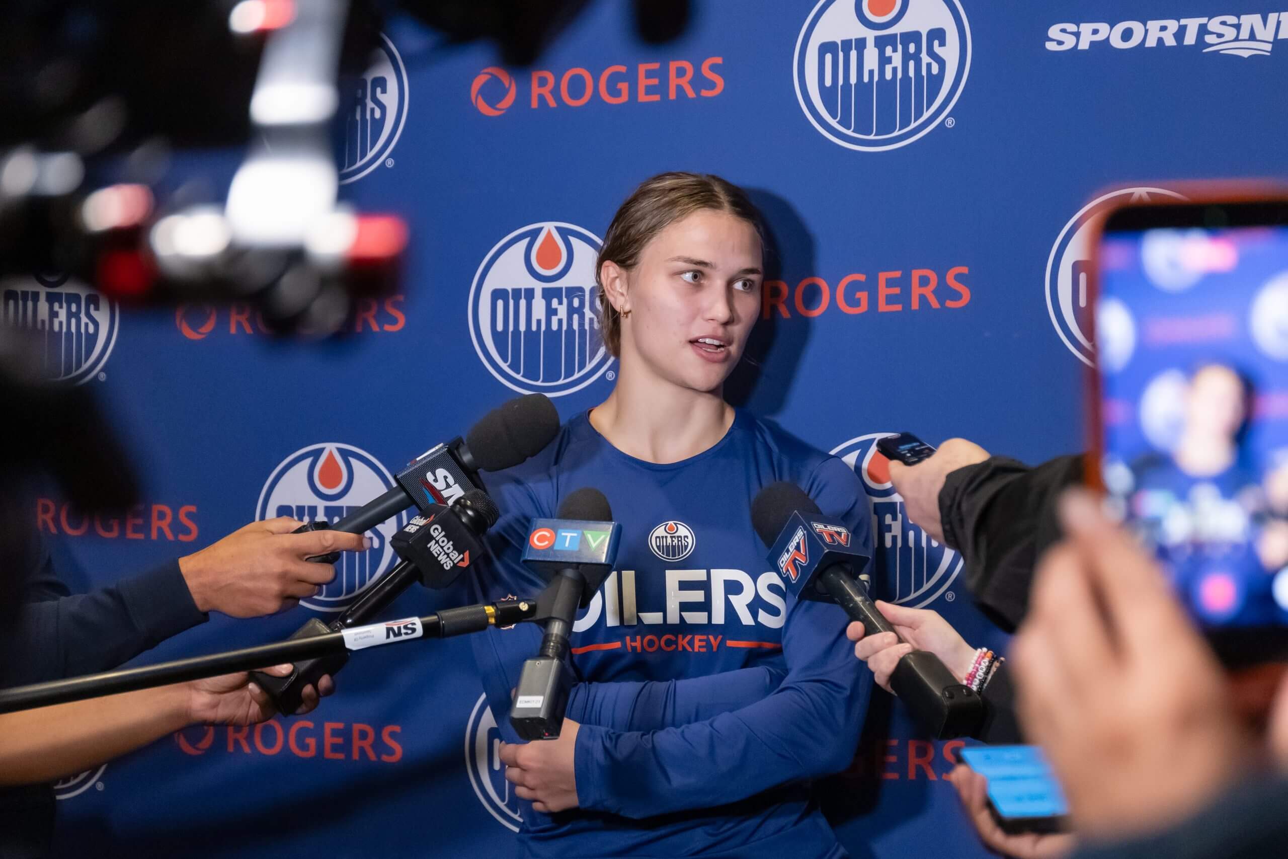 Caitlin Kraemer speaks into several microphones and two phones in front of an Oilers backdrop.