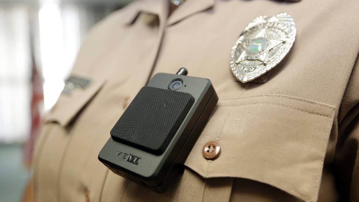 A close-up of a police officer's uniform shows a body-worn camera clipped to the front of the shirt beneath a law enforcement badge.