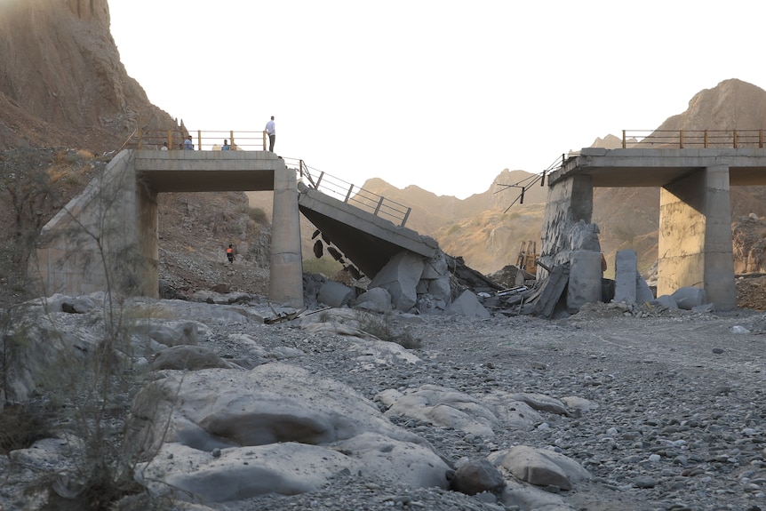 A landscape image shows a broken down concrete bridge and a few people standing on the edge of it to look down