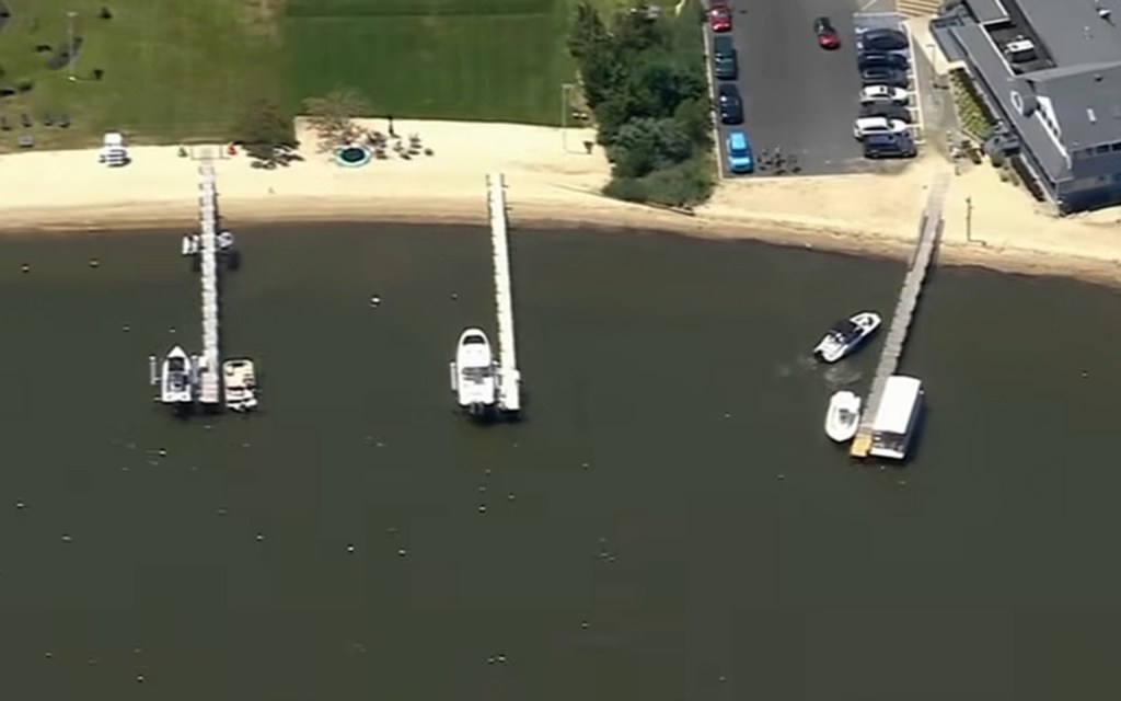 Aerial view of multiple piers with docked boats along a shoreline with sandy beaches, grassy areas, a road with parked cars, and buildings.
