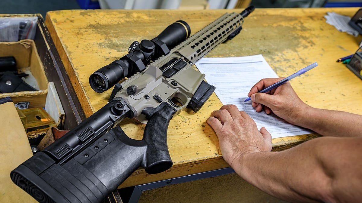 Male buyer signs paperwork beside an AR-15 rifle in a gun shop.