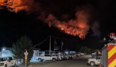 A diagonal line of fire burns on a distant mountainside in the night, beyond an illuminated line of trucks.