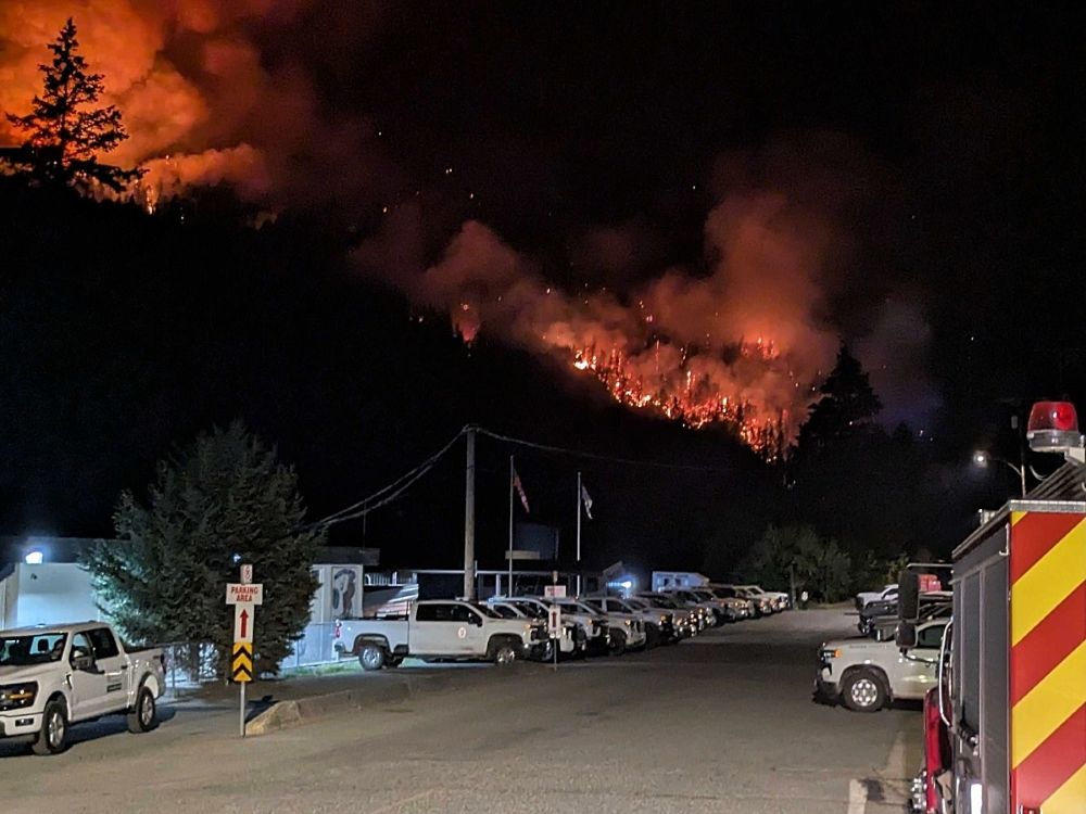 A diagonal line of fire burns on a distant mountainside in the night, beyond an illuminated line of trucks.