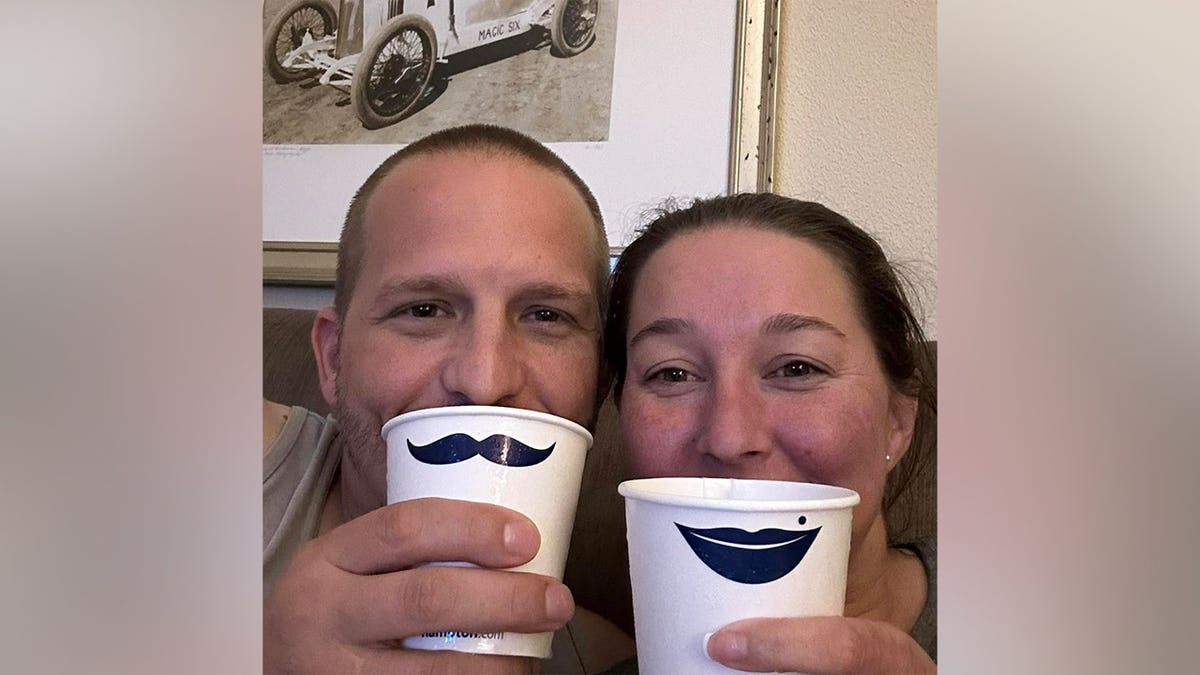 Justin and Lena Elrod smiling closely together while holding paper cups printed with a mustache and lips design.