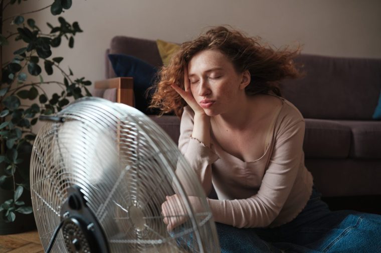 Suffering from summer heat at home, exhausted young woman sitting near electric fan enjoying fresh air, cooling down during heatwave - stock photo