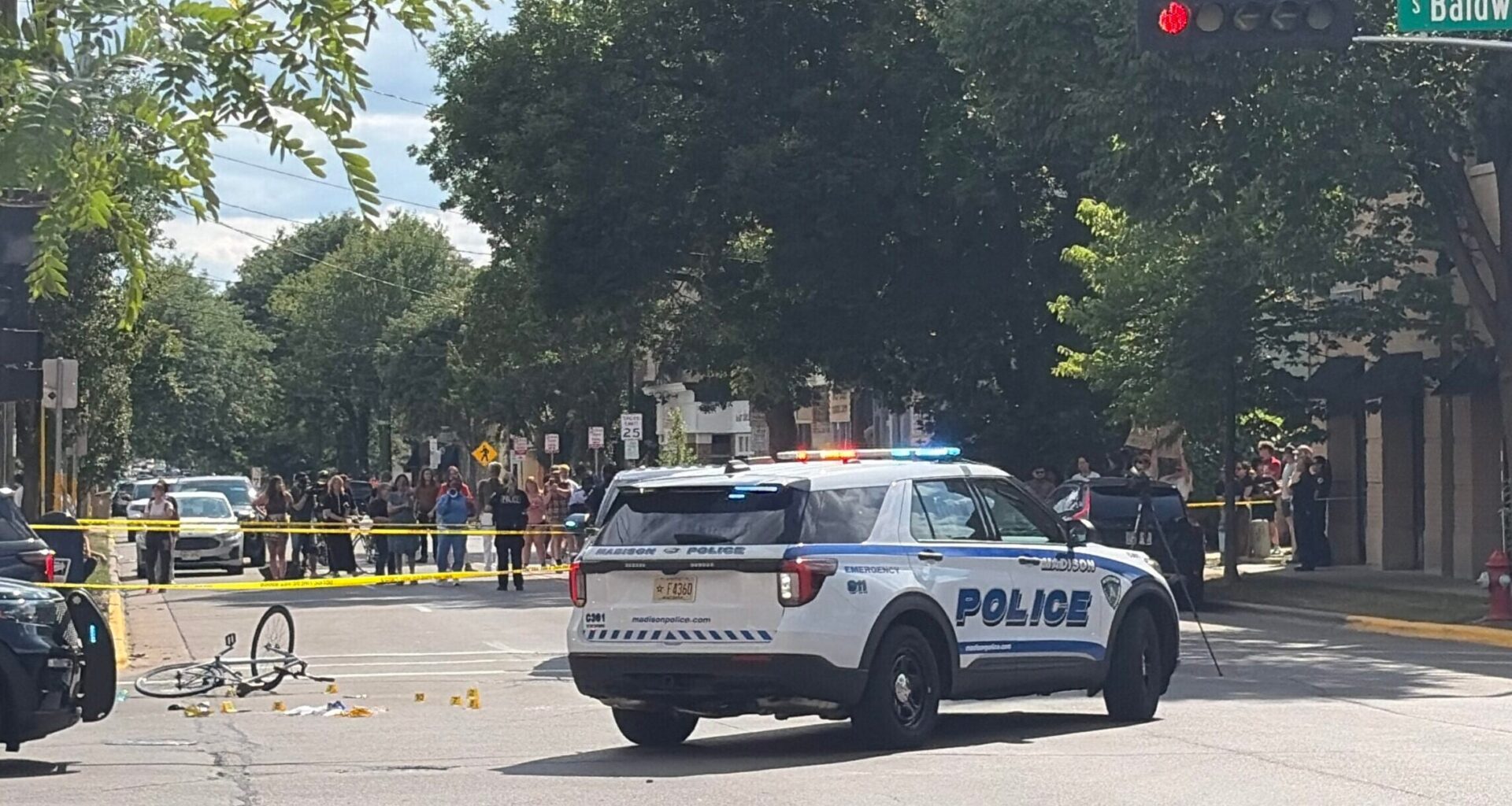 A police SUV blocks an intersection with yellow tape, a bicycle on the ground, traffic cones, and a crowd gathered nearby under trees.