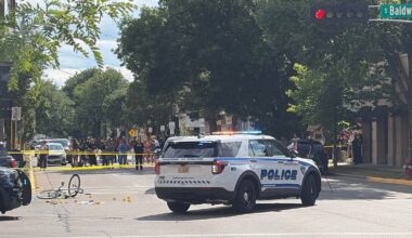 A police SUV blocks an intersection with yellow tape, a bicycle on the ground, traffic cones, and a crowd gathered nearby under trees.