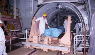 Workers in protective suits handle large blue cylindrical containers, moving them on a rail-mounted platform inside an industrial facility.