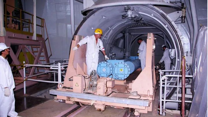 Workers in protective suits handle large blue cylindrical containers, moving them on a rail-mounted platform inside an industrial facility.