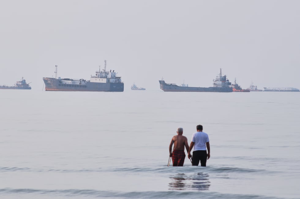 Two men wade in the waters of the Strait of Hormuz with vessels anchored in the background,...