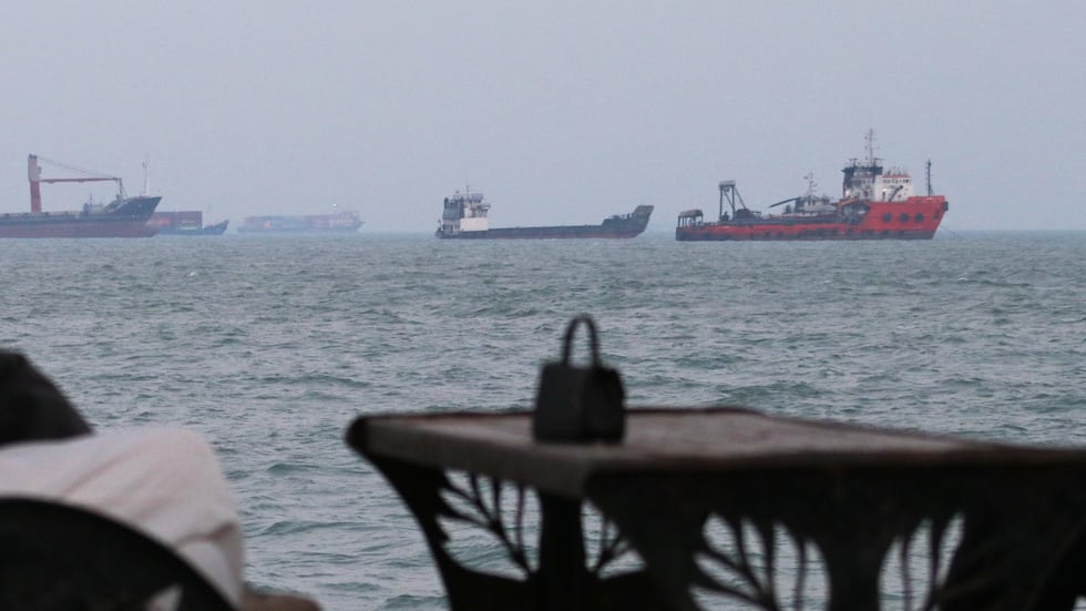 Residents check their cellphones as they sit at a cafe overlooking commercial vessels anchored...