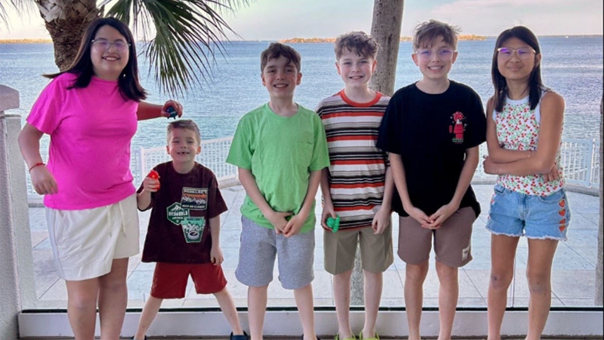 Six children, ranging from a young boy to teenagers, stand in a row smiling on a waterfront patio with a palm tree and calm water behind them.