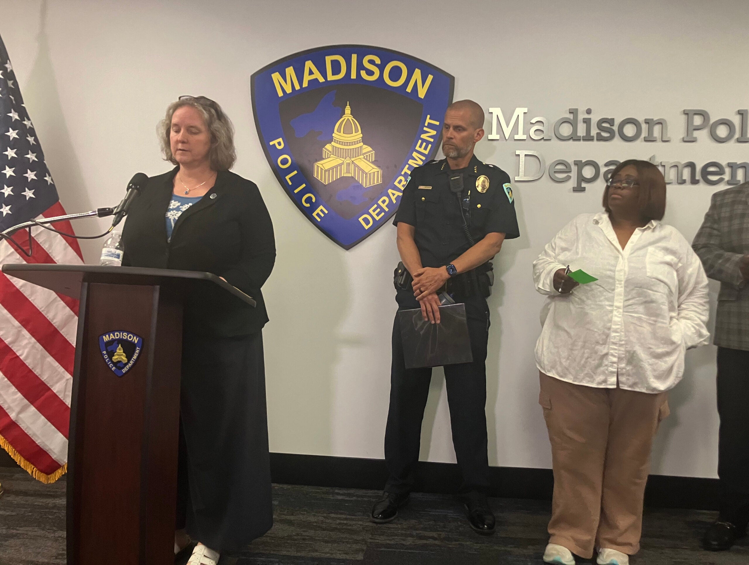 A woman speaks at a podium with the Madison Police Department logo behind her. A police officer and two other people stand nearby.