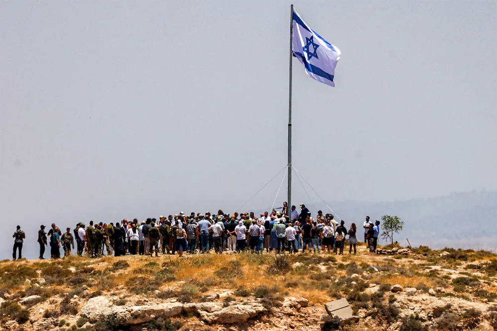 A large crowd is gathered at the top of a dry, rocky hill beneath a tall flagpole flying an Israeli flag against a clear sky.