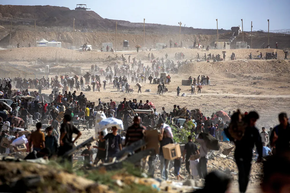 A wide, high-angle shot shows a large crowd of people walking across a dusty and dry landscape while carrying bags and belongings. Dirt mounds and arid hills line the background.
