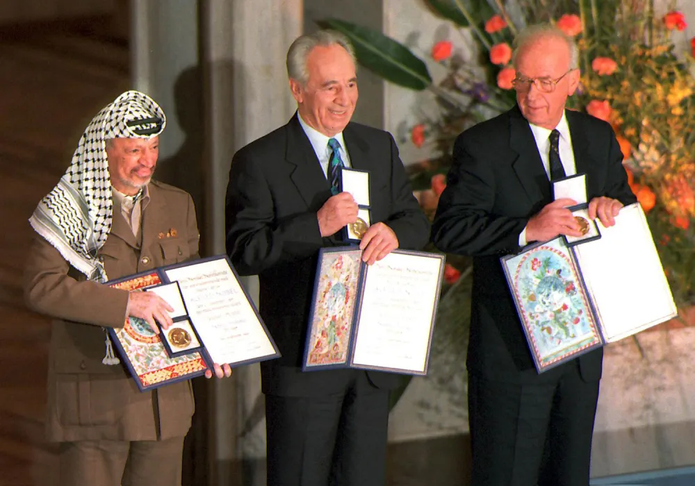 Three men in formal attire stand side by side, each holding a framed diploma and a medal in a small box. The man on the left wears a keffiyeh and tan jacket, while the two men next to him wear dark business suits and ties.