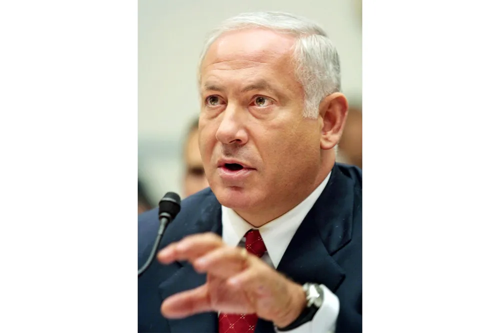A close-up shot of Benjamin Netanyahu wearing a dark suit and red tie and speaking while gesturing forward with his hand near a microphone.