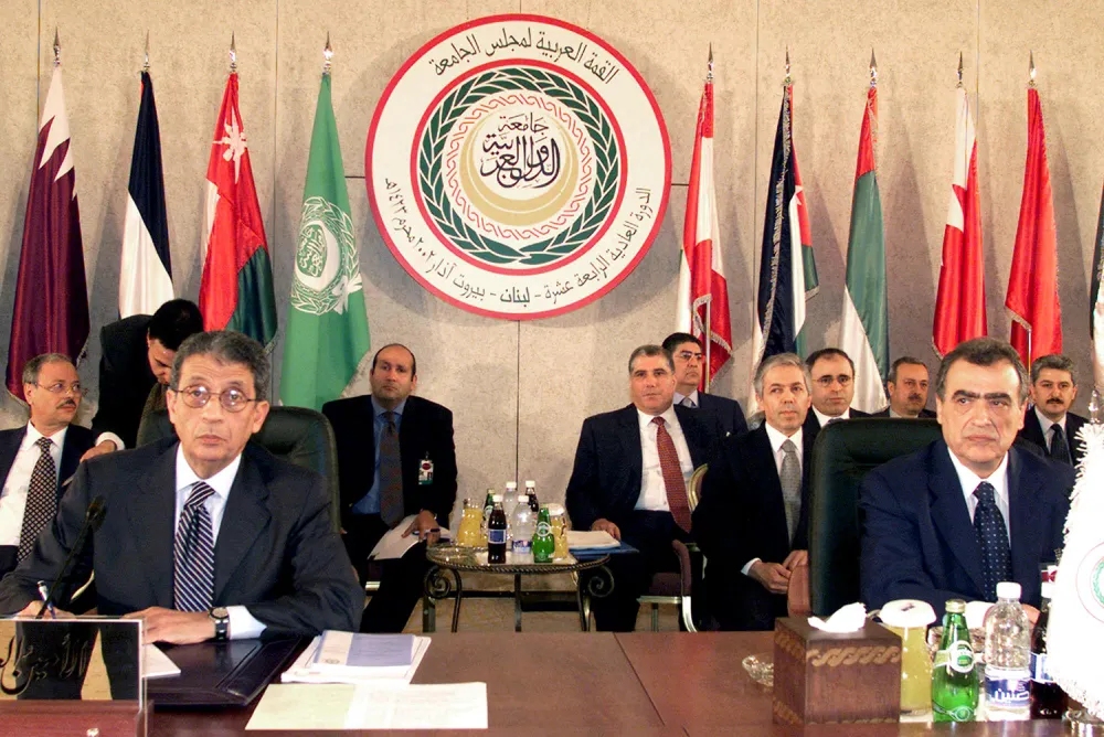 Men in suits sit at a long wooden table during a formal meeting. Behind them, a large circular emblem with Arabic script is centered on the wall, flanked by several international flags.