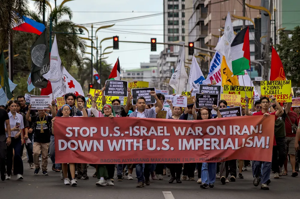 A crowd of demonstrators marches down an urban street holding flags and carrying a large red banner that reads "STOP U.S.-ISRAEL WAR ON IRAN! DOWN WITH U.S. IMPERIALISM!"