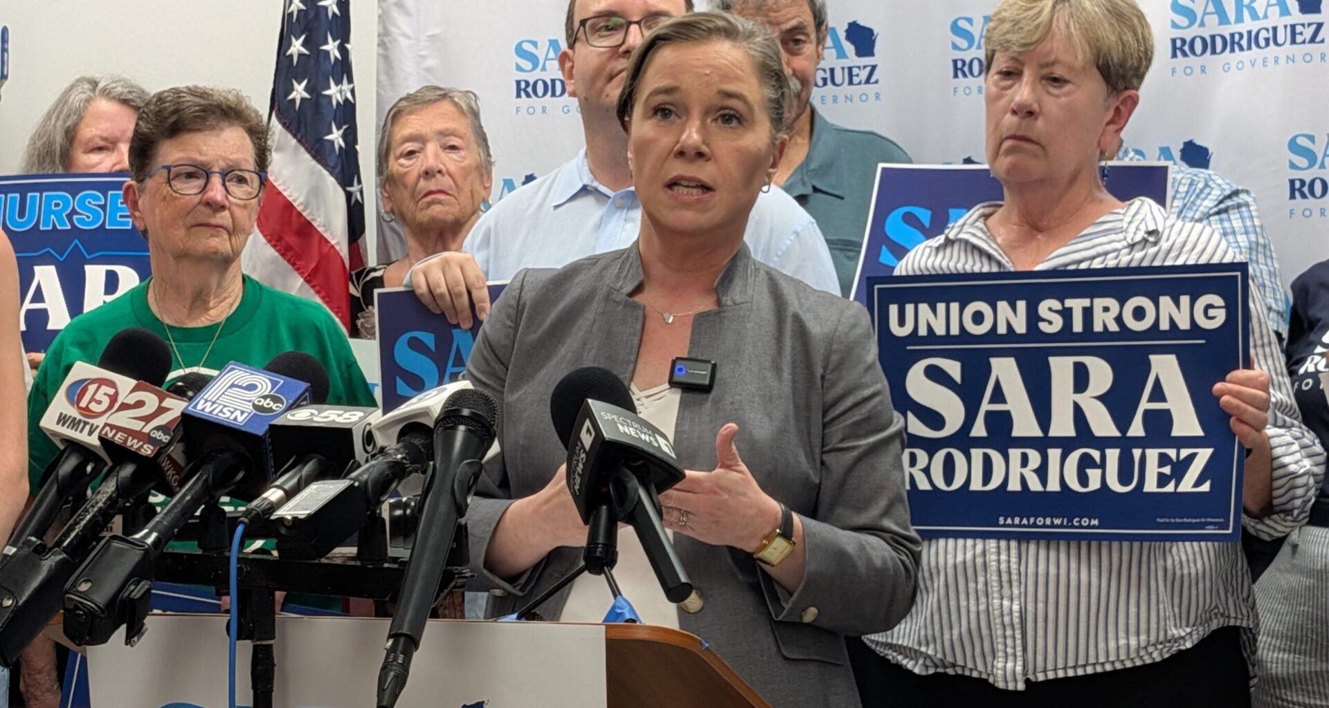 A woman speaks at a podium surrounded by supporters holding “Sara Rodriguez” signs, microphones in front, American flag and campaign banners visible in the background.