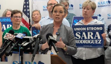 A woman speaks at a podium surrounded by supporters holding “Sara Rodriguez” signs, microphones in front, American flag and campaign banners visible in the background.