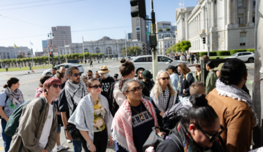 A diverse group of people wearing sunglasses and keffiyeh scarves waits together on a city sidewalk near an intersection with historic buildings in the background.