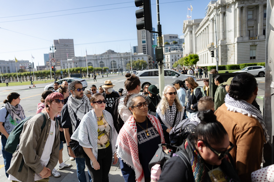 A diverse group of people wearing sunglasses and keffiyeh scarves waits together on a city sidewalk near an intersection with historic buildings in the background.
