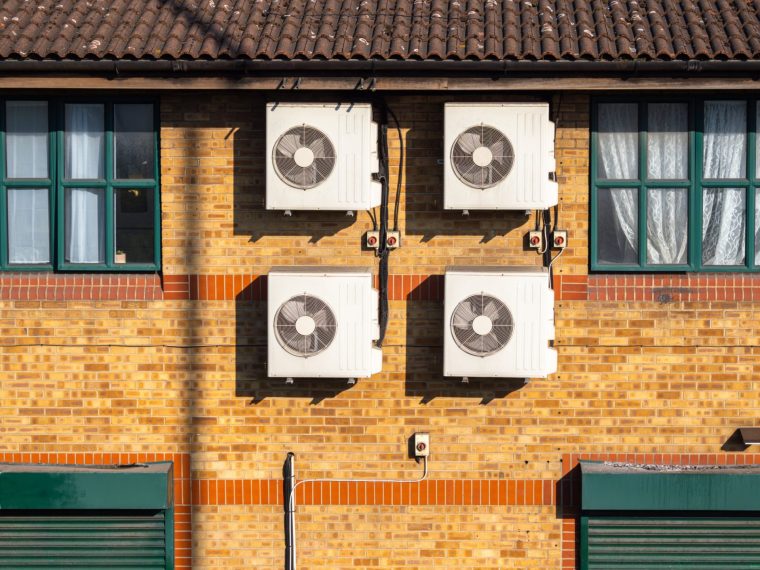 A close-up view of four air conditioning units mounted on a brick wall of a building. The units are white and rectangular, contrasting with the warm tones of the brick. Windows with green frames.