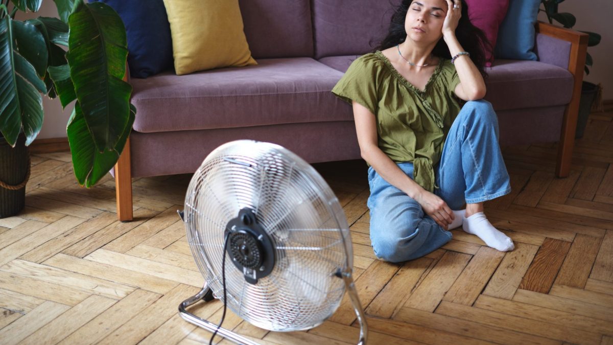 Tired exhausted young woman sitting on floor in front of electric fan, cooling down refreshing during heatwave. Summer heat, Air Conditioner Problems
