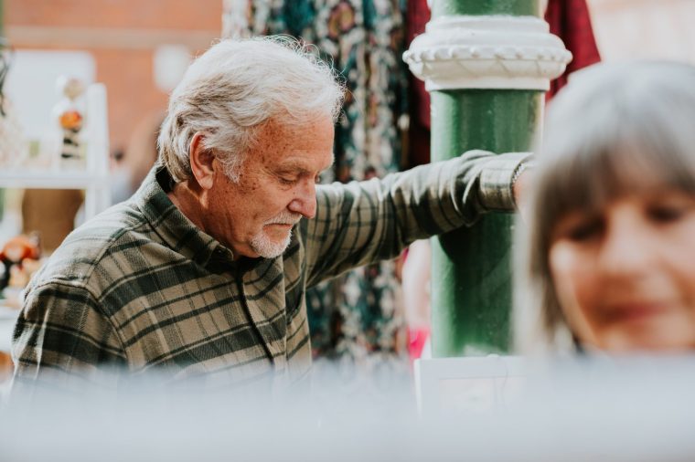 A senior couple spend time in an indoor sunny market. The woman looks through a clothes on a rail as her husband waits patiently behind her.