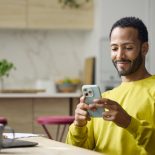 Smiling young freelancer using smart phone. Male entrepreneur is surfing on internet. Man is working from home.