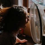 A little girl sits at a table indoors on a hot day. Her hair blows around uncontrollably as the old fashioned large desk fan blows in her face.